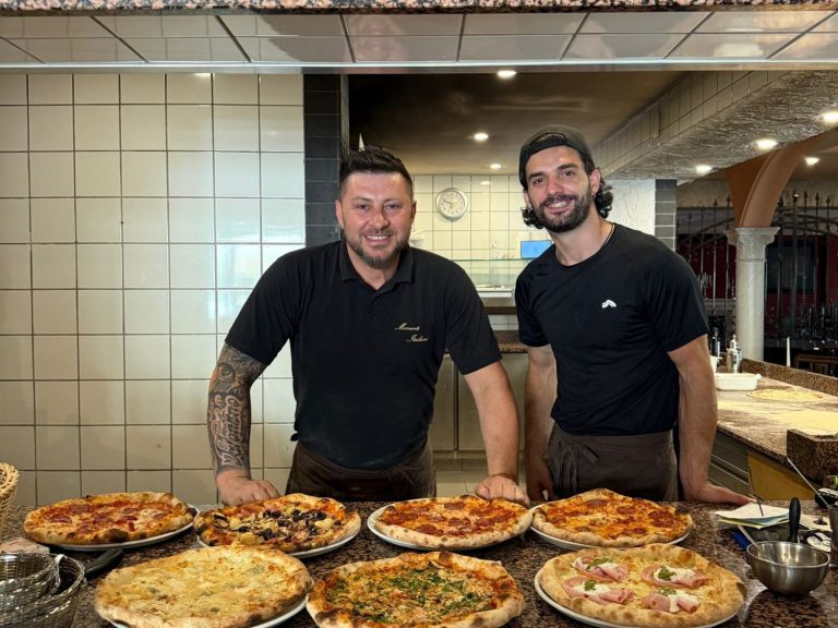 Father & Son Zwei Männer in schwarz, lächelnd, neben verschiedenen Pizzen in einem Restaurant.