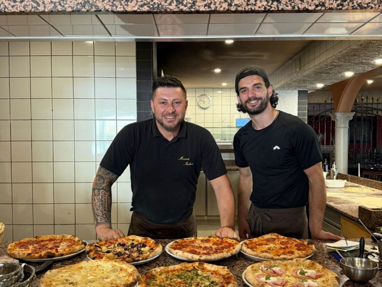 Father & Son Zwei Männer in schwarz, lächelnd, neben verschiedenen Pizzen in einem Restaurant.