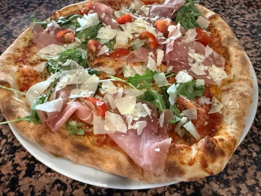 Pizza mit Schinken, Rucola, Kirschtomaten und Parmesan auf einem Teller.